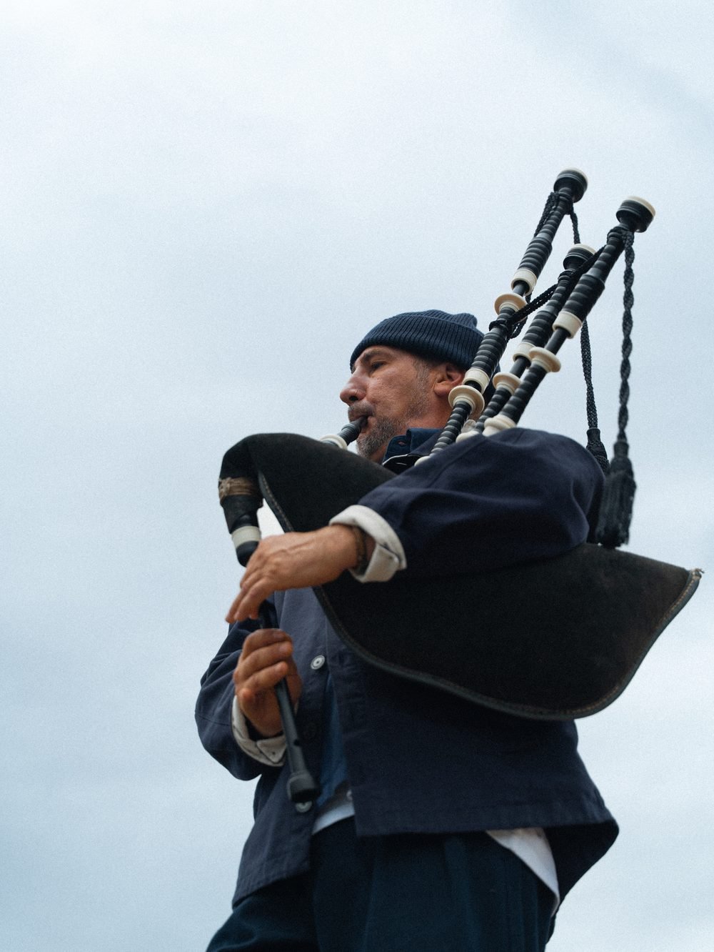 Sonneur de cornemuse de profil, bonnet et veste bleu marine, face à la mer.