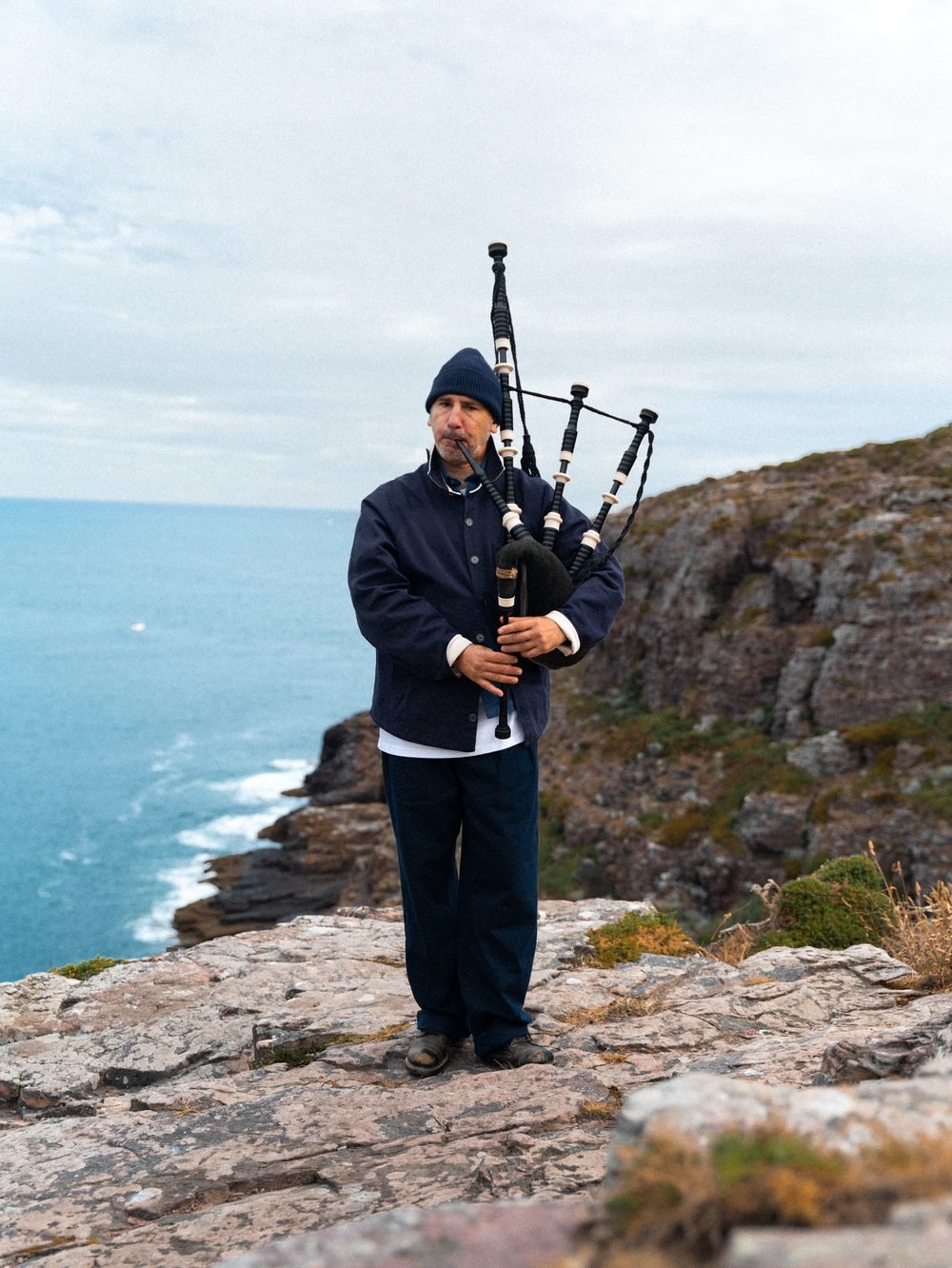 Le sonneur debout sur la lande rocheuse, la cornemuse sur l'épaule, la mer derrière lui.