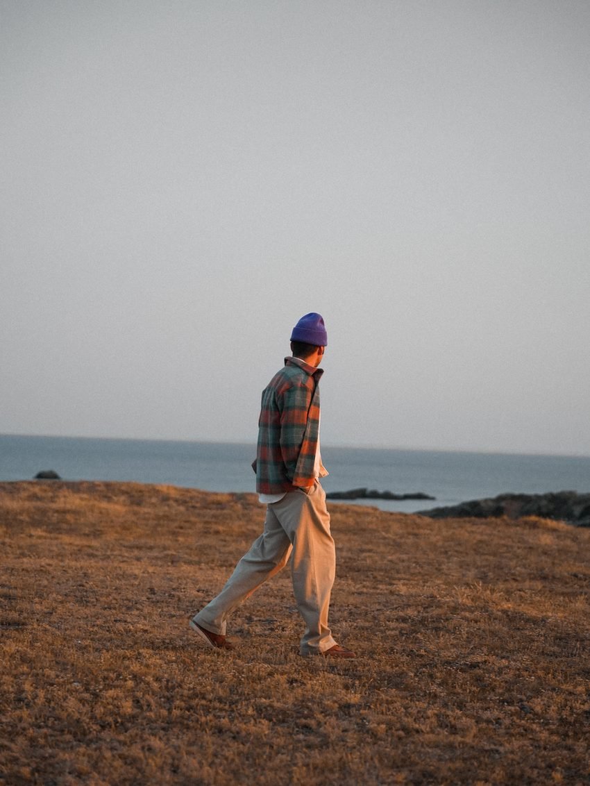 Homme marchant sur la lande rase au bord de l'eau, bonnet violet et surchemise à carreaux.