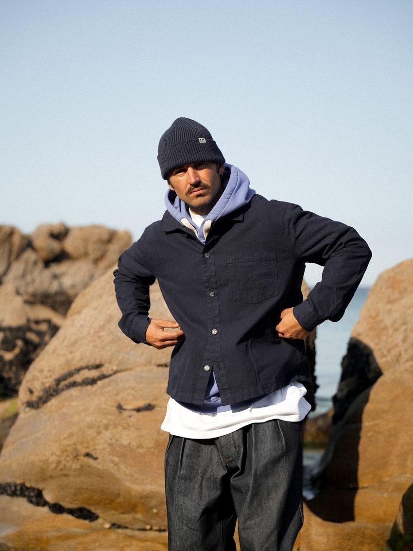 Homme debout sur les rochers, veste bleu marine, capuche claire et bonnet.