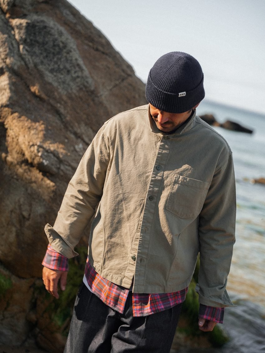 Homme en surchemise beige et bonnet bleu marine, adossé aux rochers.