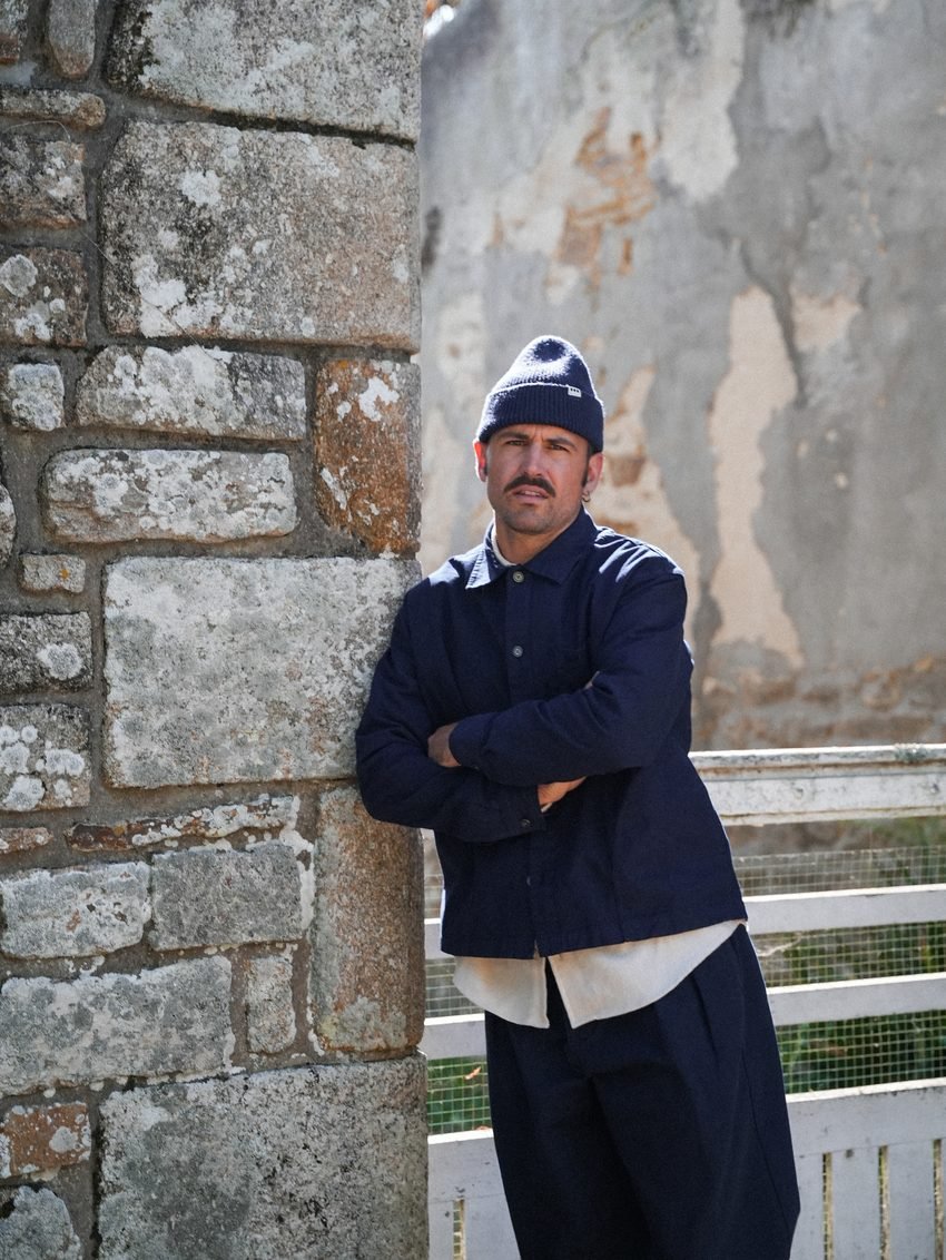Homme en veste bleu marine adossé à un mur de pierre, bras croisés.