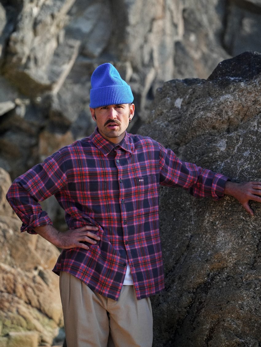 Homme en chemise à carreaux rouges et bonnet bleu vif, appuyé à un rocher.