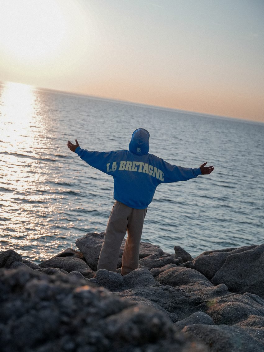 Homme de dos, bras écartés face au coucher de soleil, sweat « LA BRETAGNE ».
