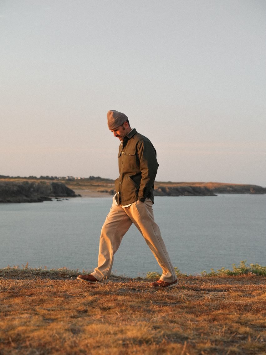 Homme marchant sur la lande en veste kaki et pantalon beige, l'eau derrière lui.
