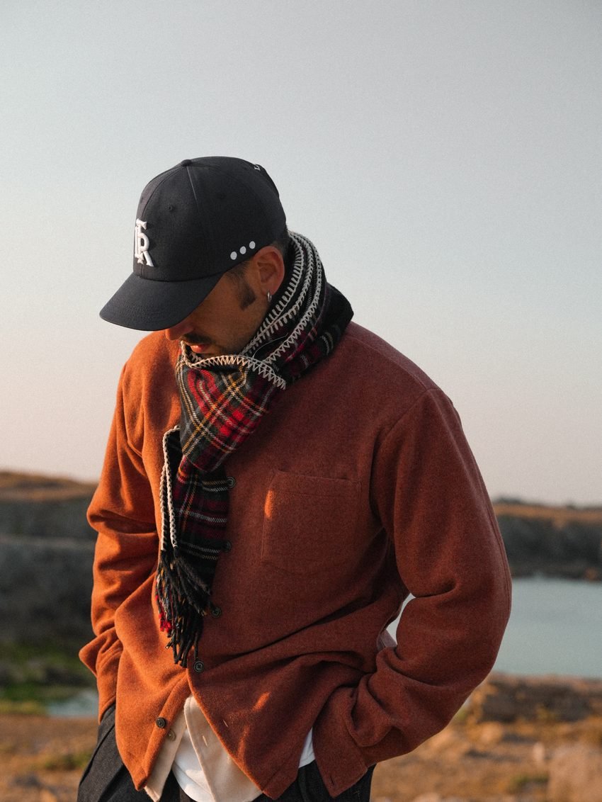 Homme en polaire orange, écharpe à carreaux et casquette, regard baissé.