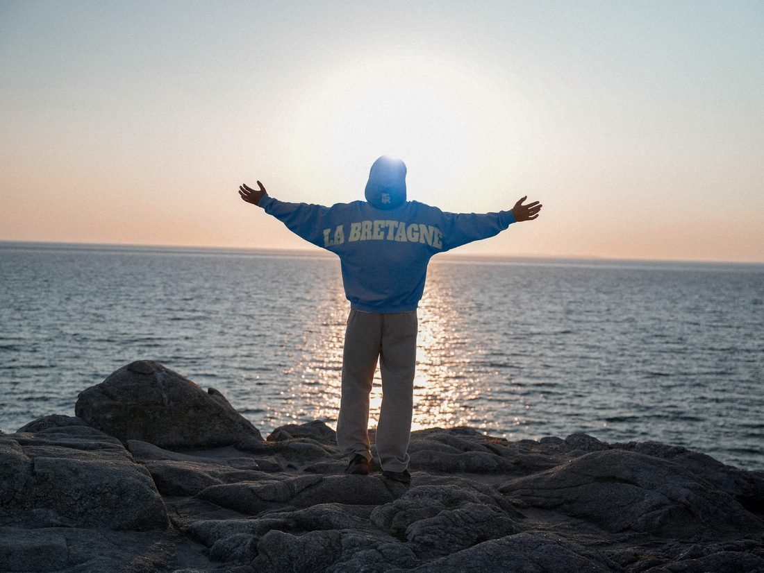 Homme de dos, bras écartés face au soleil couchant, sweat « LA BRETAGNE ».