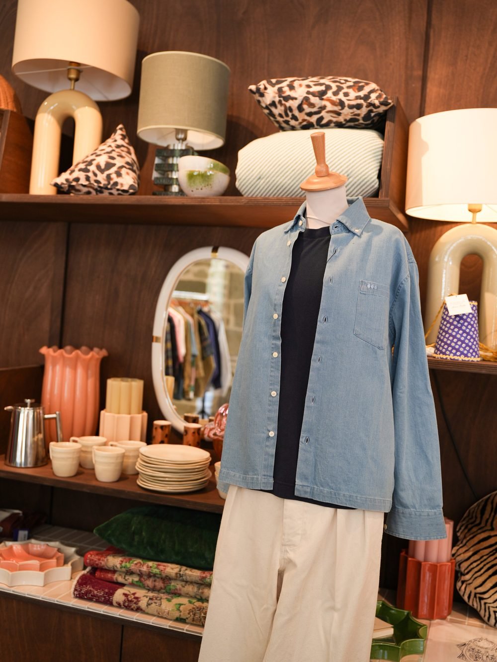 Mannequin en chemise en jean et pantalon beige, devant des étagères de lampes et de vaisselle.