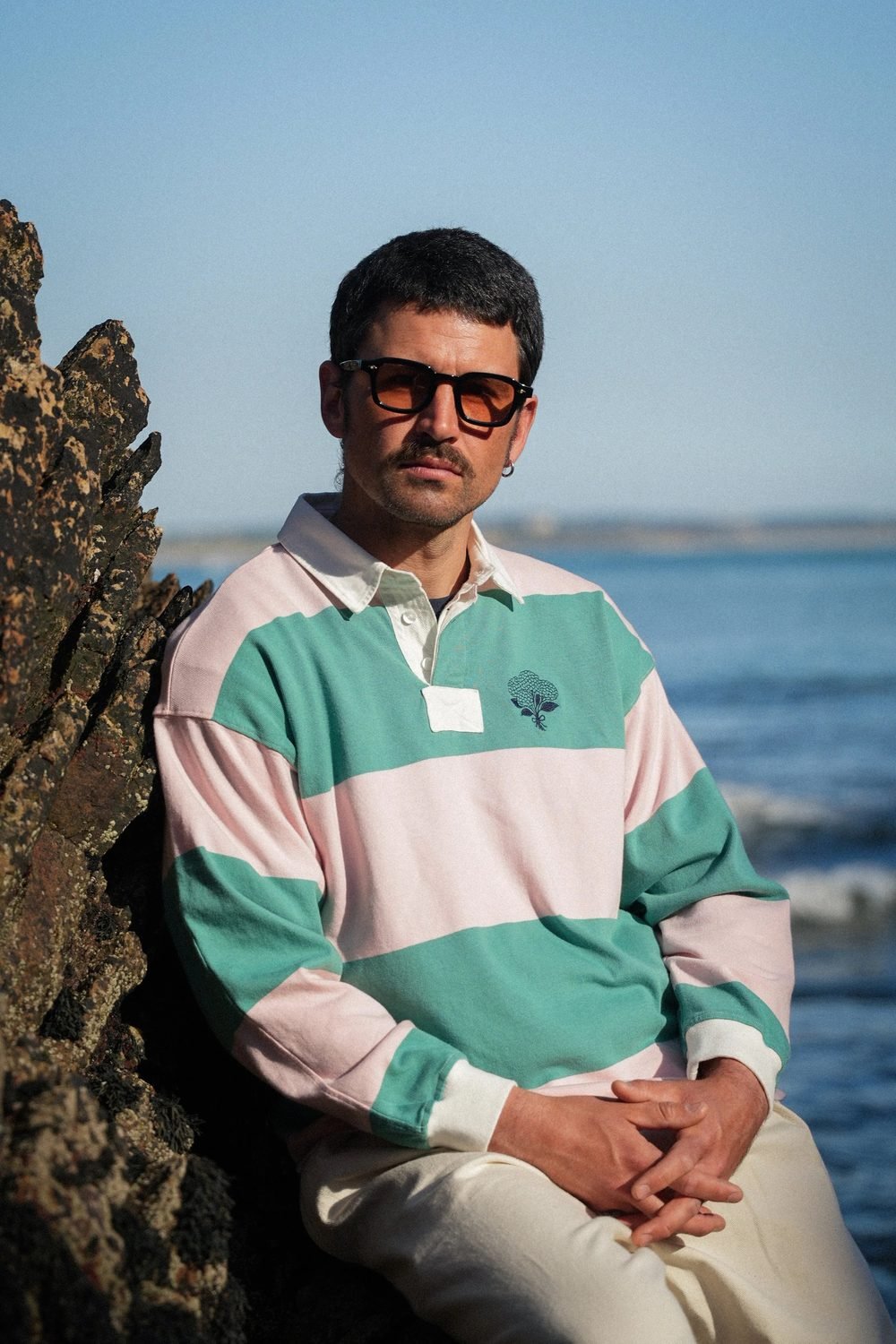 Homme assis contre un rocher en polo de rugby rayé rose et vert, la mer en arrière-plan.