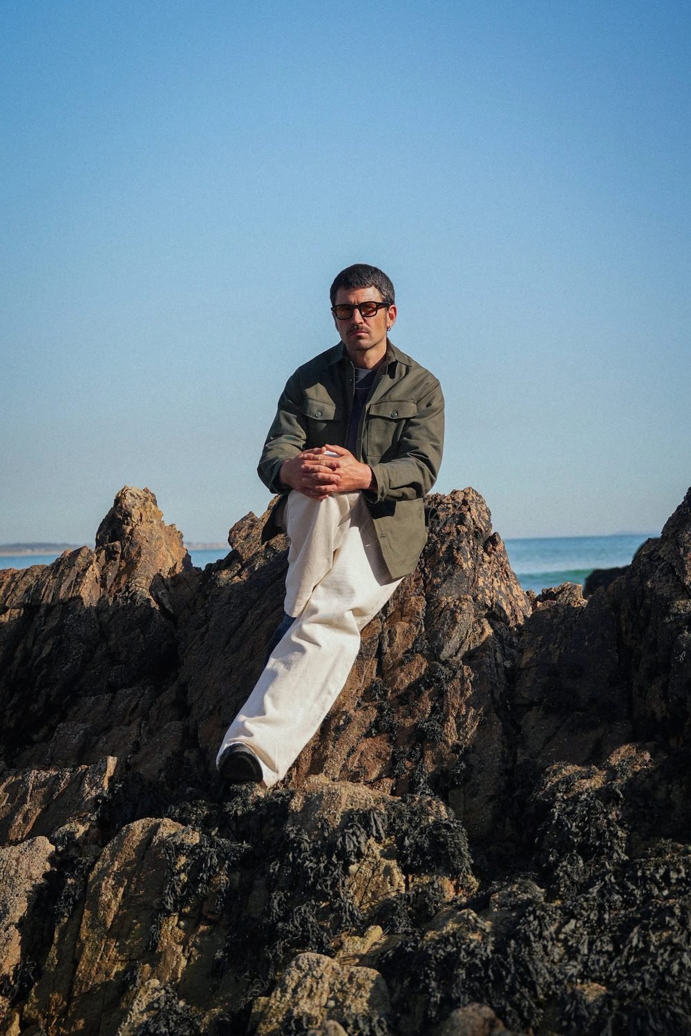 Homme assis sur les rochers en veste kaki et pantalon blanc, face à l'océan.