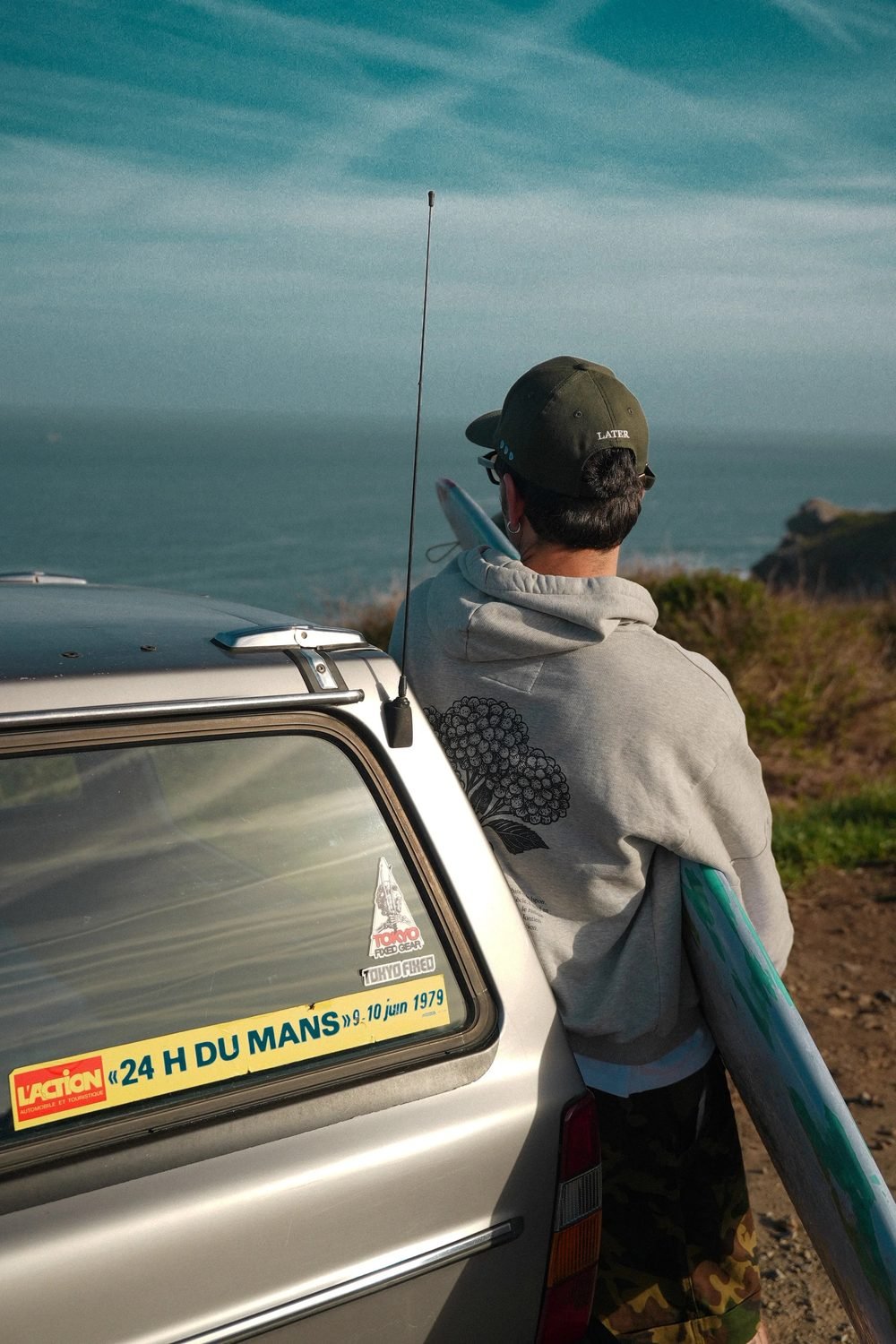 Homme de dos adossé à une voiture ancienne au bord de la côte, sweat à capuche gris à motif floral.