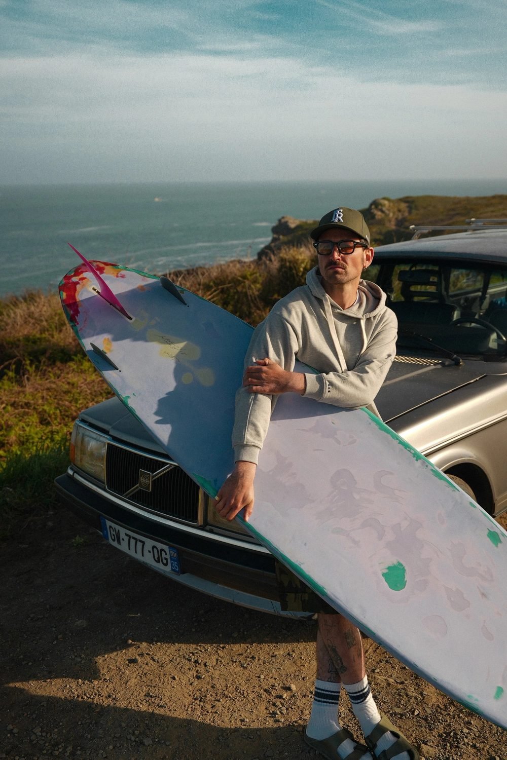 Homme portant une planche de surf devant une voiture ancienne, casquette et sweat à capuche.