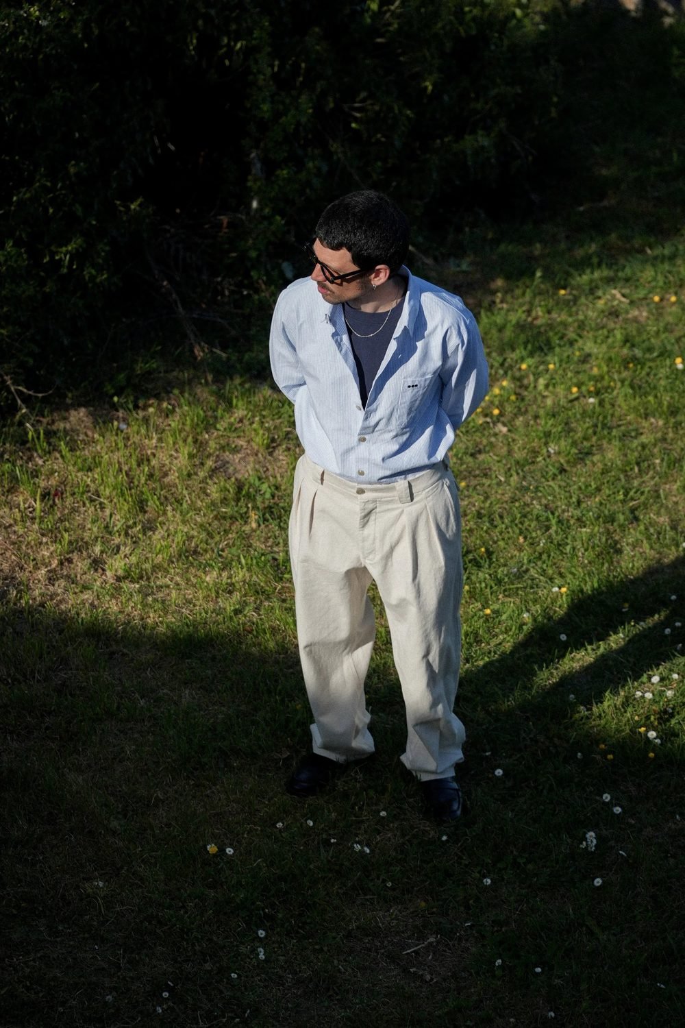 Homme debout dans l'herbe, chemise claire ouverte et pantalon beige.